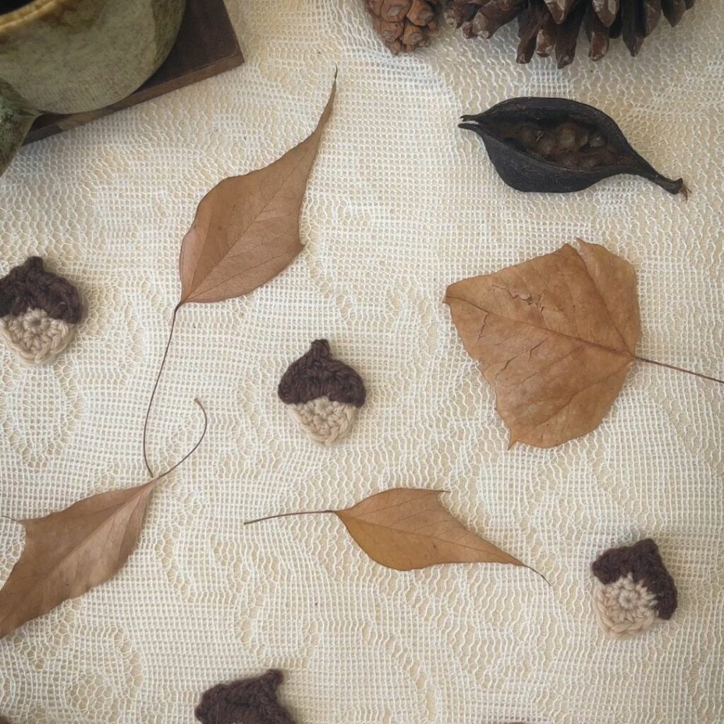 Crochet acorns on a cream lace background surrounded by leaves with pinecones and a cup of coffee.