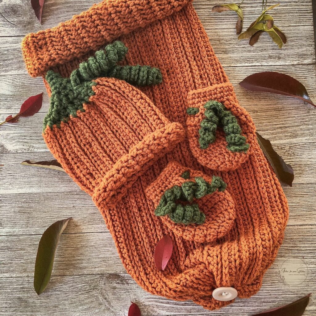 Vintage-style photo of a handmade crochet pumpkin baby set, including a ribbed cocoon sack with button closure, matching hat with green stem detail, and two booties, all styled on a rustic wooden background with scattered autumn leaves.