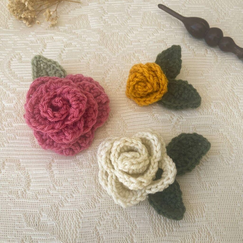 Crochet rose and leaf set featuring roses in raspberry, gold, and off white with green leaves, displayed on a lace background beside a wooden crochet hook
